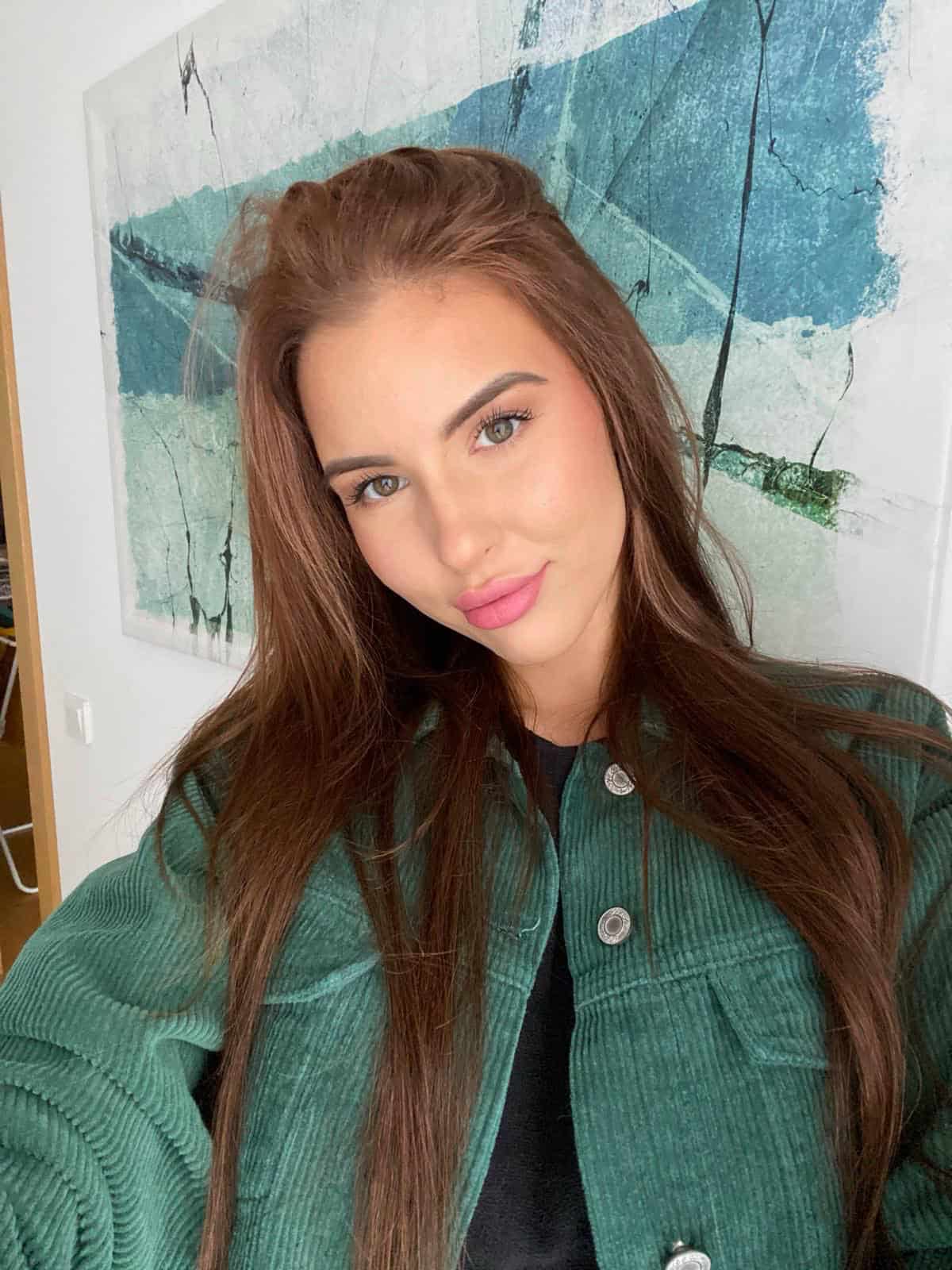 Sieviete ar brūnu matu sakopojumu, zaļu džinsu jaku un mākslas darbu fonā, moderns portrets.